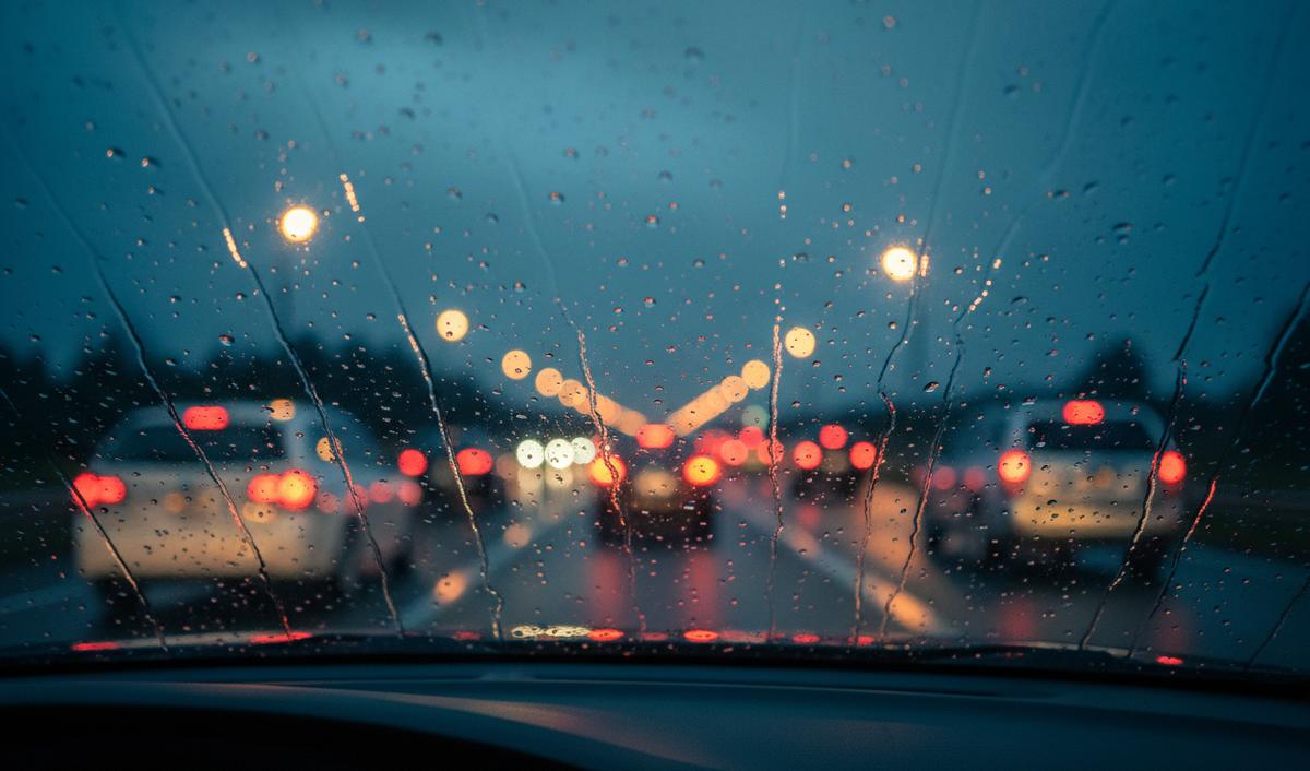 Rain on a car windshield at dusk, with blurred red tail lights and orange street lights on the highway ahead